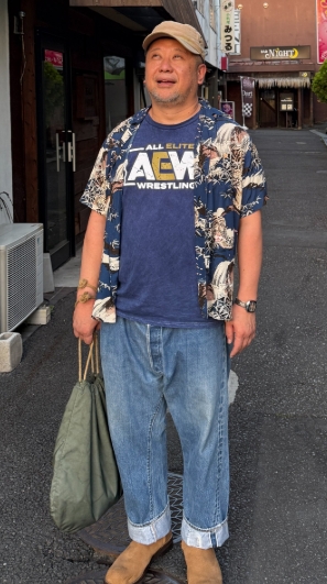 ケンドーコバヤシが街中でカジュアルな私服姿を披露。AEWロゴ入りTシャツに花柄シャツを重ね、デニムとスエードブーツを合わせた個性的なファッション。
