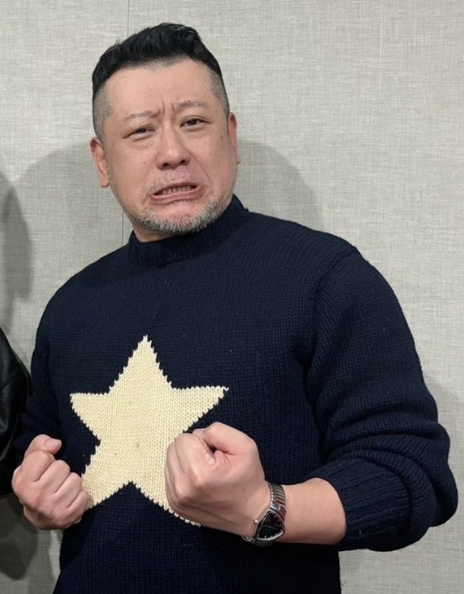 ケンドーコバヤシが星柄ニットを着用し、拳を握って力強いポーズをとる姿。短髪とヒゲが特徴的な表情豊かなショット。