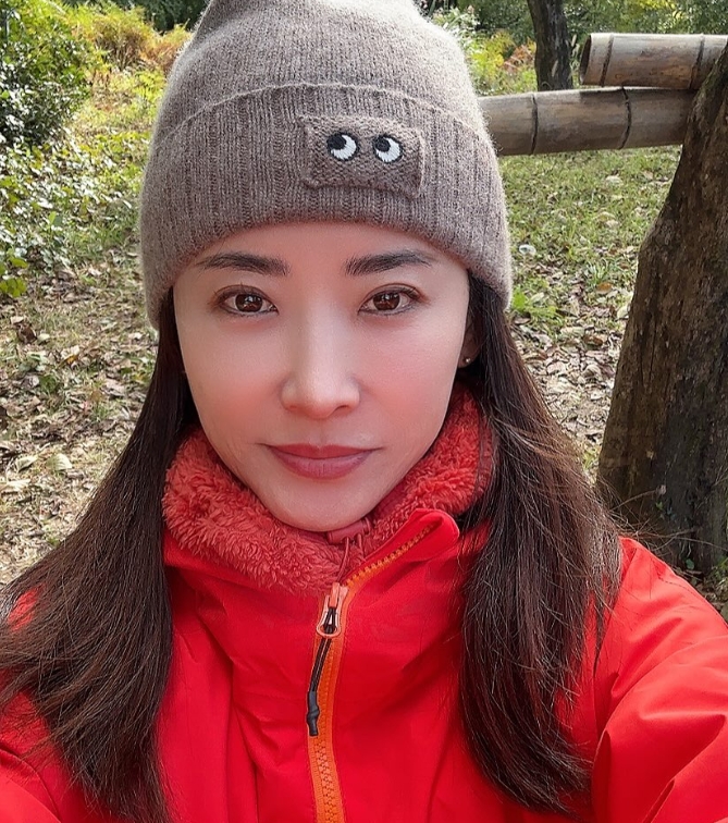 参政党から衆議院選挙に出馬した島村かおる氏が、自然豊かな屋外で赤いジャケットと茶色のニット帽を着用している様子。