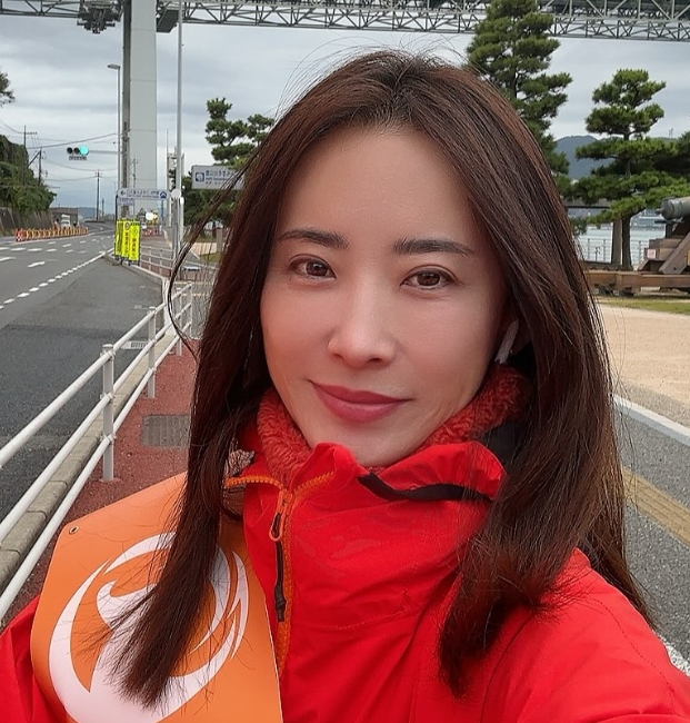 参政党から出馬した島村かおる氏が屋外で自撮りする様子。赤いジャケットとオレンジのたすきを着用し、背景には日本の道路標識と交通インフラが見える。